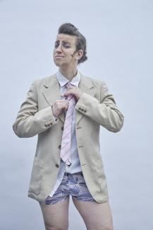 Drag King character Richard P. Dick Van Johnson wears a tan suit jacket, no pants, and a matching pink tie and underwear. He's fixing his tie.