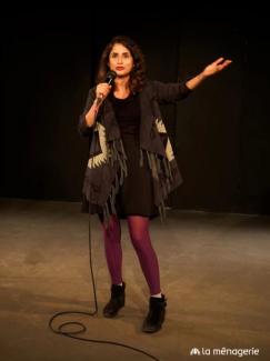 Carmen speaks into the microphone on a wide stage and raises her hands. She wears red tights, red lipstick and a poncho.