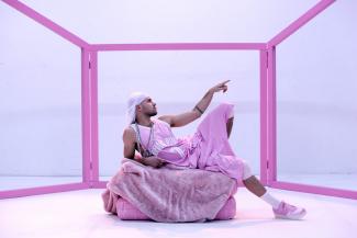 Ein Performer, der bekleidet ist mit einem weiten ärmellosen Trikot, einer rosafarbenen Sporthose und einem weißen Bandana, liegt auf einem rosafarbenen Polster. Sein linker Zeigefinger zeigt in die Ferne, sein linkes Bein ist angewinkelt. Die Pose ist Michelangelos Deckenfresko "Die Erschaffung Adams" nachempfunden. Im Hintergrund wird das Bühnenbild umrahmt von einem Gerüst aus rosafarbenen Streben. 