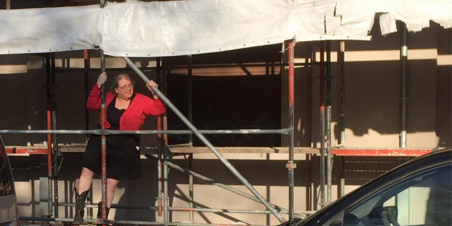 A white woman wearing a short black dress, coral cardigan sweater, black cowboy boots, and glasses is climbing up some scaffolding in front of a building. She is looking out to her left and smiling slightly, or maybe she is squinting in the afternoon soon. The edge of a white tattered tarp flaps behind her.