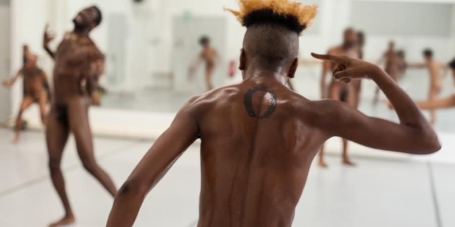 Ein nackter Oberkörper eines Tänzers ist im Vodergrund von hinten zu sehen, auf dem Rücken trägt er ein Tattoo mit einem Kreis, er zeigt mit seinem ausgestreckten Zeigefinger auf seinen Kopf, er schwitzt. Im Hintergrund sind noch mehr Tänzer:innen zu sehen, alle sind nackt.  