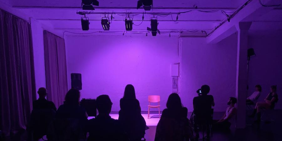 Aufnahme einer in Violett beleuchteten Bühne. Die Bühne, die hinter dem Publikum zu sehen ist, ist leer.